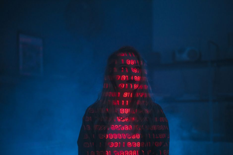 A person stands in a dimly lit environment shrouded in blue and red lighting, with the person's face and upper body obscured by a hooded garment displaying red binary code projections. The background 