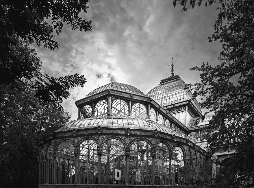 A large, historic glass-domed conservatory with classical architectural details, situated beside a reflecting pond. The building features a prominent central dome with a decorative spire, flanked by s